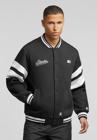 Black varsity jacket with white stripes on the sleeves, ribbed neckline, snap front closure, and an embroidered logo. Soft fabric texture.