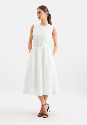 Vrouw met bruin haar in een paardenstaart, gekleed in een witte mouwloze midi-jurk en zwarte sandalen met hoge hakken, staand met handen in de zakken.