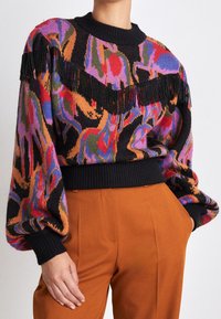 Pull en tricot avec une base noire ornée de motifs abstraits colorés en rose, vert et bleu, agrémenté d'une frange noire sur la poitrine.