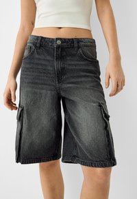 Personne portant un short cargo en denim noir délavé et un débardeur blanc court, debout devant un fond clair uni.