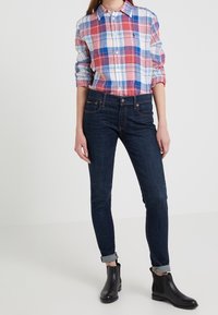 Camicia a quadri rossa, blu e bianca abbinata a jeans skinny blu scuro. Stivali neri alla caviglia con tacco basso. Maniche arrotolate visibili.