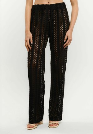 Czarne spodnie na szydełku o szerokich nogawkach, noszone z beżowymi, otwartymi sandałami na obcasie, prezentowane na osobie stojącej na białym tle.