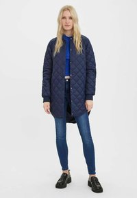 Granatowa pikowana długa kurtka z okrągłym dekoltem, napami i ściągaczami. Noszona na niebieskiej bluzce i ciemnych dżinsach, w zestawie z czarnymi loafersami.