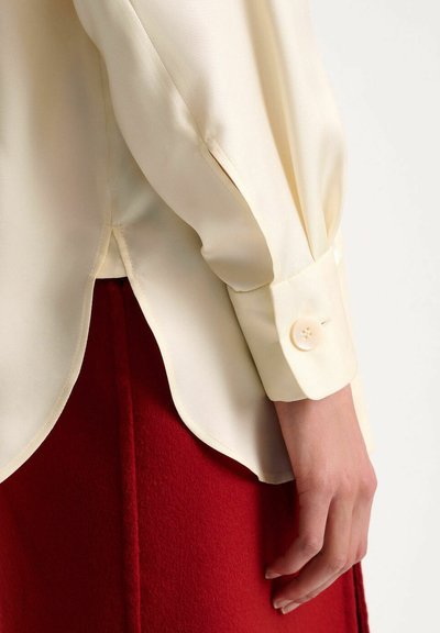 Blusa crema con orlo rotondo, maniche lunghe con polsini a bottone, abbinata a una gonna rossa scura con dettaglio della cucitura centrale.