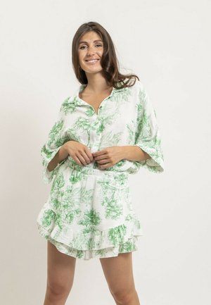 Femme souriante aux longs cheveux bruns portant un chemisier boutonné à imprimé floral blanc et vert, assorti d'un short à volants.
