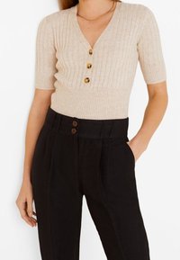 Haut en tricot côtelé beige à manches courtes et col en V, comportant trois boutons ronds marron. Associé à un pantalon noir taille haute avec deux boutons.