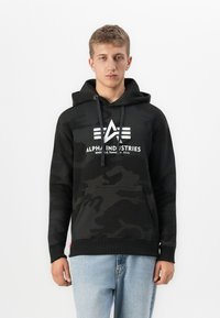 Joven vestido con una sudadera con capucha de camuflaje negro con el logo de Alpha Industries y unos vaqueros azul claro, de pie frente a un fondo blanco liso.
