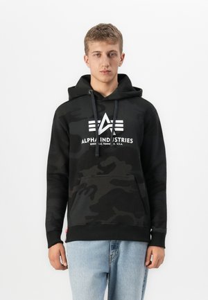 Joven vestido con una sudadera con capucha de camuflaje negro con el logo de Alpha Industries y unos vaqueros azul claro, de pie frente a un fondo blanco liso.
