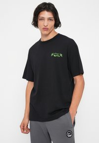 Jauns vīrietis ar plecu garuma matiem, ģērbies melnā Nike SB T-kreklā un pelēkā Nike treniņbiksēs, stāv ar vienu roku kabatā.
