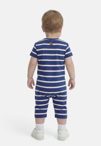 Νήπιο που στέκεται με την πλάτη γυρισμένη, φορώντας μπλε-άσπρη ριγέ φορμάκι με ένα μικρό αρκουδάκι στην πάνω πλάτη, λευκές κάλτσες και λευκά αθλητικά παπούτσια.