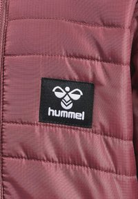 Burgundowa pikowana kurtka z czarno-białą naszywką logo, gładka w dotyku, z zapięciem na zamek. Wypełniona w celu zapewnienia ciepła, z poziomym przeszyciem.