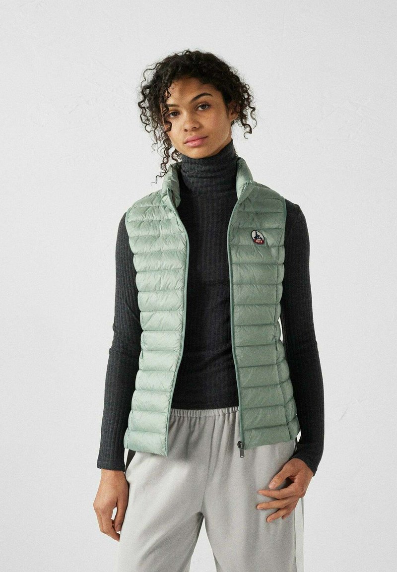 Gilet matelassé vert menthe avec un quilt horizontal, col haut et fermeture éclair à l'avant, superposé sur un col roulé noir à côtes et un pantalon gris.