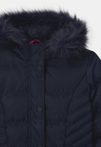 Giacca puffer navy con cappuccio in pelliccia sintetica, caratterizzata da un design trapuntato, bottoni a pressione neri e un'etichetta rosa all'interno.