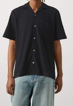 Mann trägt ein schwarzes kurzärmeliges Hemd mit Brusttasche und hellblauen Jeans vor einem einfarbigen Hintergrund.