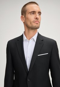 Schwarzer Herrenblazer mit Reverskragen, Ein-Knopf-Verschluss und einem weißen Einstecktuch über einem weißen Hemd mit kleinen blauen Punkten.