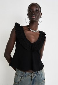 Black cotton top with a deep V-neck and intricate eyelet ruffle details along the neckline. Paired with light blue denim jeans.