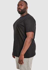 T-shirt nera a maniche corte con collo rotondo, realizzata in cotone. Il modello è in posizione laterale, indossa pantaloni corti grigi. Design semplice senza motivi.