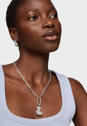 Collar de cadena de plata con un colgante de oso de peluche texturizado, llevado sobre una camiseta sin mangas de canalé azul claro. Pendientes de aro simples completan el look.