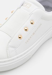 Weißer Sneaker mit glattem Lederfinish, elastischem Band, goldenen Nieten und dezent eingraviertem Branding. Verfügt über eine strukturierte Gummisohle.