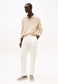Beige stripete langermet polo skjorte, hvite avslappede bukser med klaffelomme, og grå slip-on sko med strukturert overflate.