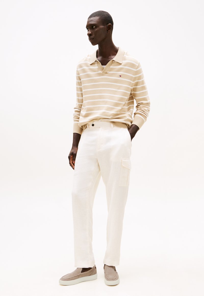 Camisa polo de manga larga a rayas beige, pantalones blancos de corte relajado con un bolsillo de solapa y zapatos grises tipo slip-on con una superficie texturizada.