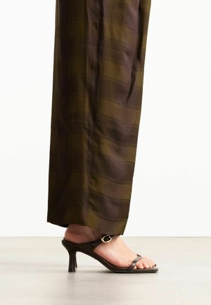 Person trägt braune karierte weite Hosen und dunkelbraune Riemchen-Sandaletten mit goldener Schnalle auf einem hellen Boden.