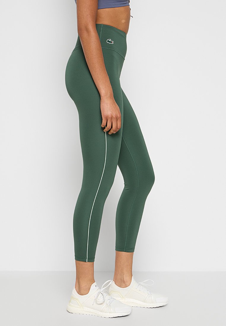 Leggings verdi con vita alta, texture liscia, striscia laterale bianca e piccolo logo sulla cintura, abbinati a scarpe da ginnastica bianche.