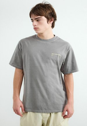 Mladý muž s hnedými vlasmi, oblečený v šedej oversize tričku s malou textovou potlačou na hrudi a svetlo béžových nohaviciach, stojaci pred bielym pozadím.