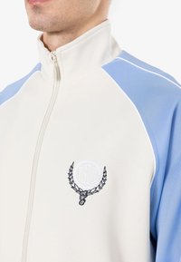 Viegls zip-up jaciņa ar krēmkrāsas ķermeni un ziliem plecu paneļiem, izšūtu logotipu un baltu apdari apkārt rāvējslēdzim un šuvēm.