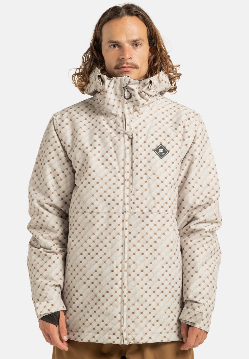 Mężczyzna w beżowej pikowanej kurtce z małym brązowym wzorem w diamenty i kapturem, stojący na jednolitym tle.