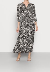 Robe longue noire et blanche à fleurs avec un décolleté en V, des manches trois-quarts et une silhouette fluide, assortie à des sandales à talons de couleur claire.