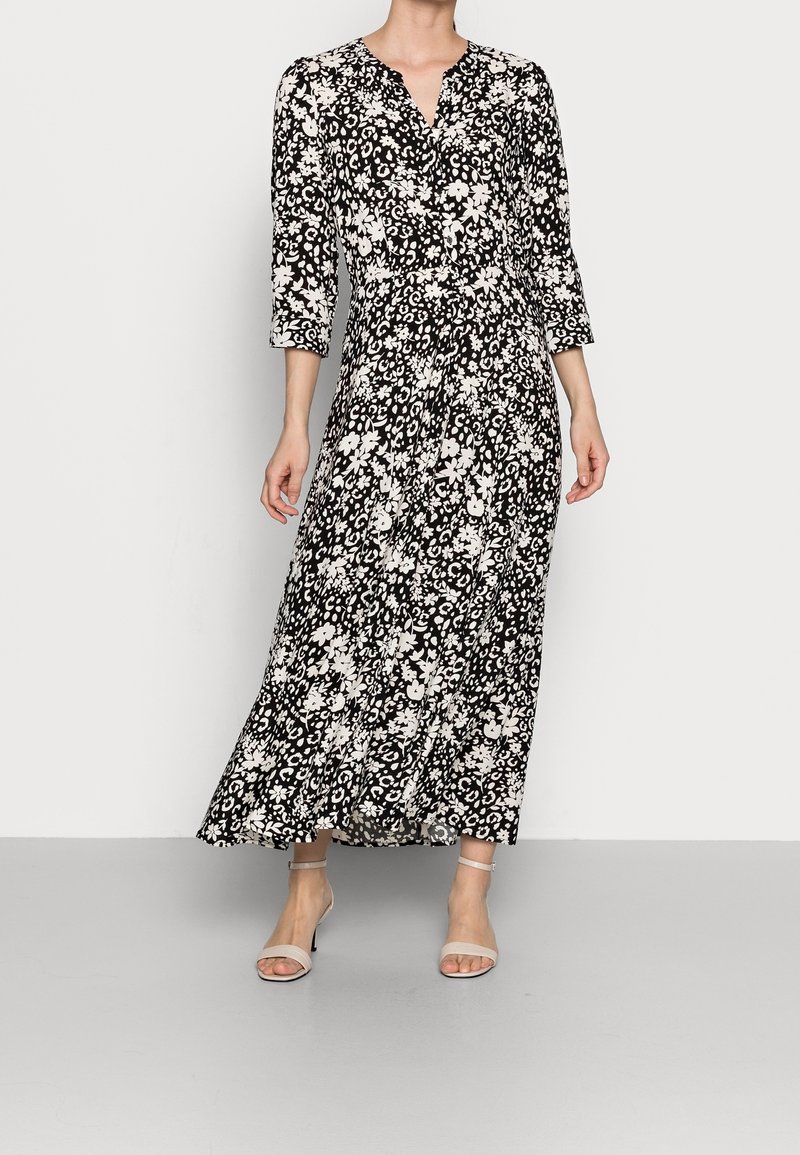 Robe longue noire et blanche à fleurs avec un décolleté en V, des manches trois-quarts et une silhouette fluide, assortie à des sandales à talons de couleur claire.