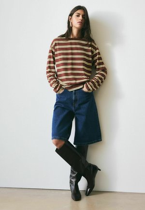 Kvinde stående op ad væg iført brun og cremefarvet stribet langærmet skjorte, denimshorts til knæet og sorte støvler til knæet.