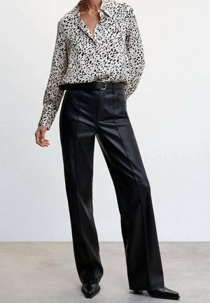 Femme portant un pantalon large en cuir noir taille haute avec une ceinture noire et un chemisier blanc à boutons avec des taches abstraites noires.