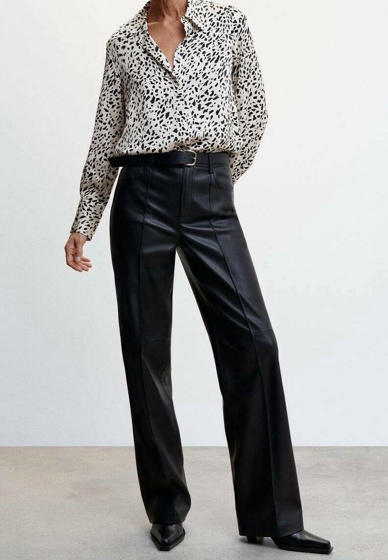 Femme portant un pantalon large en cuir noir taille haute avec une ceinture noire et un chemisier blanc à boutons avec des taches abstraites noires.
