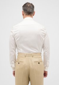 Weißes Langarmhemd mit spitzem Kragen, kombiniert mit beigen Hosen mit zwei Gesäßtaschen und Knopfdetails.