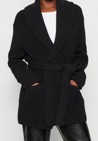 Cardigan nero lavorato a maglia con ampio colletto a scialle, maniche lunghe, cintura con nodo e due tasche frontali. Il tessuto testurizzato conferisce un aspetto accogliente.