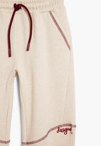 Lichtbeige sweatpants met een geribbelde tailleband, bordeauxrode trekkoord en contrasterende stiksels, met "ontworpen" geborduurd in bordeaux.