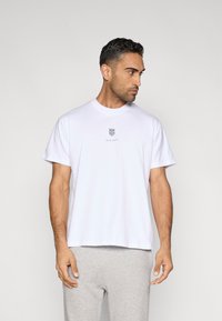 T-shirt branca de algodão com mangas curtas, gola redonda, apresentando um pequeno logótipo em forma de escudo no centro da frente, combinada com calças de fato de treino cinzentas.