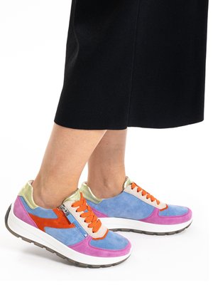 Voeten met meerkleurige suède sneakers met oranje veters en ritssluitingen aan de zijkant, gecombineerd met zwarte cropped broeken tegen een witte achtergrond.