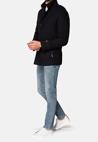 Homme portant une veste noire boutonnée, un jean bleu clair et des baskets blanches, debout les mains jointes devant un fond uni.