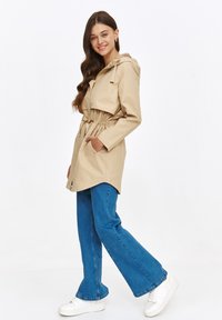 Beżowy, lekki płaszcz z kapturem i ściągaczem w talii, w połączeniu z dzwonowymi niebieskimi dżinsami i białymi trampkami. Gładki materiał, casualowy design.