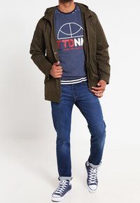 Olivgrön huva jacka med dragkedja, marinblå sweatshirt med tryckt logo, blå jeans och vita sneakers med blå detaljer.