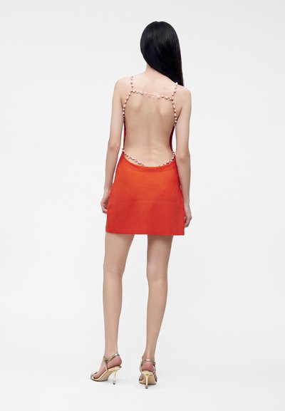 Femme aux longs cheveux noirs portant une mini-robe orange dos nu avec des bretelles en perles et des sandales dorées à talons hauts, de dos.