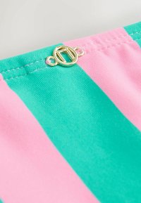 Rohelise ja roosa triibuline kangas, mille ülaserval on kuldne ringjas kinnis, nähtavate õmblusdetailidega õmblustes.