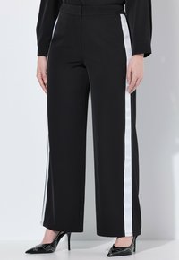 Svarta vida byxor har vita sidostråk, en slät tygtextur och ett åtsittande midjeband. De är ihop med spetsiga svarta klackar.