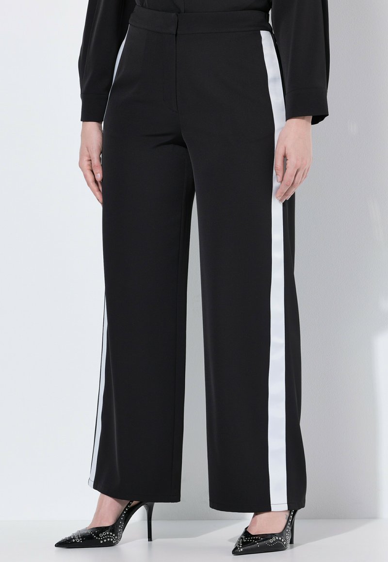 Svarta vida byxor har vita sidostråk, en slät tygtextur och ett åtsittande midjeband. De är ihop med spetsiga svarta klackar.