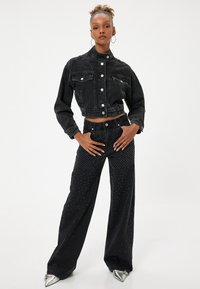 Chaqueta de mezclilla negra recortada con cierres de botón y dos bolsillos frontales, combinada con pantalones negros de tiro alto que presentan adornos de tachuelas metálicas.