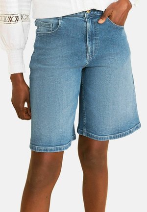 Mid-length blue denim shorts on a person with one hand in pocket, paired with a white long-sleeve top featuring lace details on the sleeves.