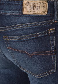 Mörkblå denimjeans med en patchetikett som läser "DIESEL." Har kontrasterande sömmar, två bakfickor och en subtil v-formad sömdesign.